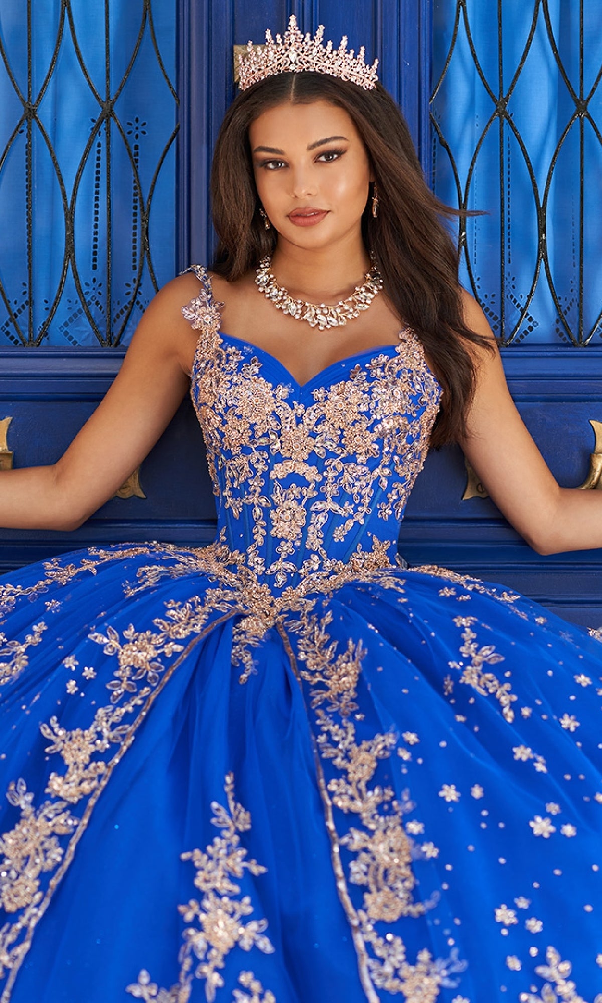 A woman wears the Quinceanera Dress PR30087 By Princesa, a royal blue gown with gold embroidery and a corset bodice, paired with a jeweled tiara, posing before ornate blue doors.